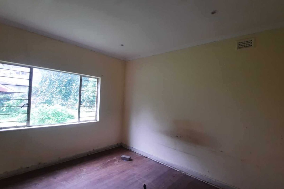 An empty bedroom featuring a steel-framed window with security bars and a ventilation grille, requiring cosmetic updates to the walls and flooring.
