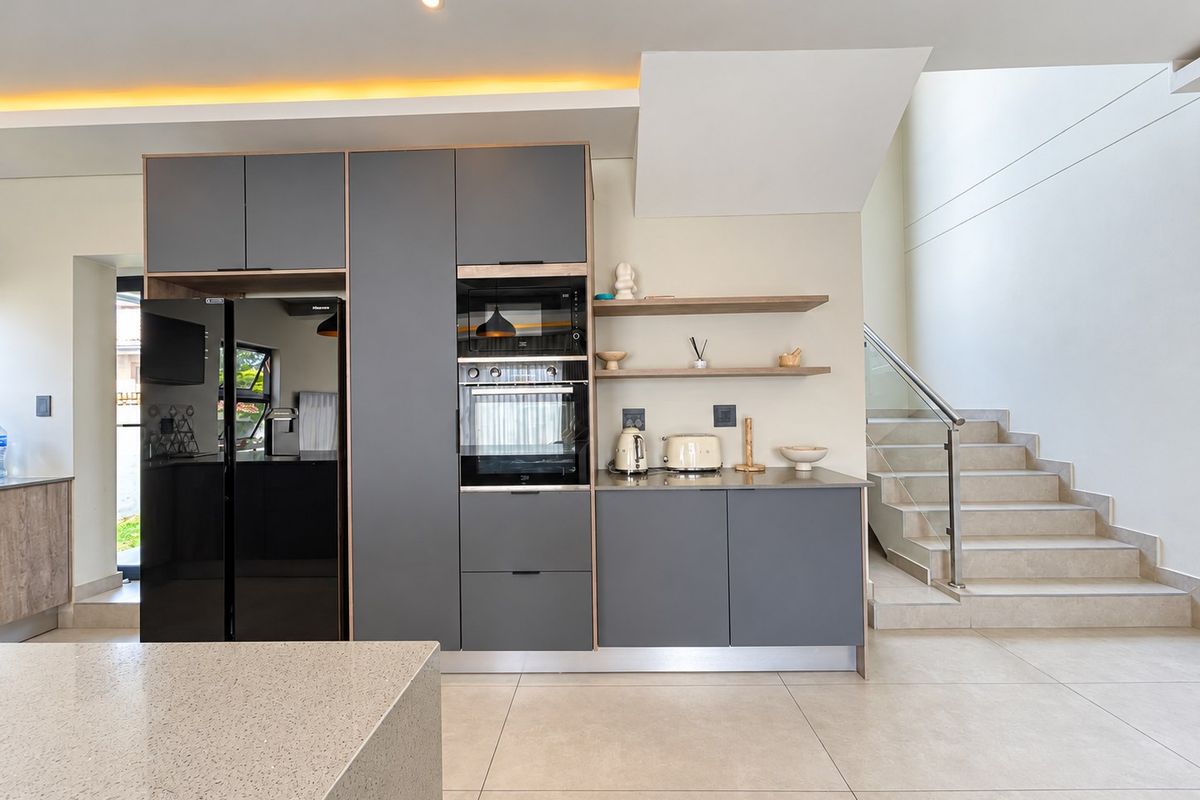 Modern kitchen featuring sleek grey cabinetry, built-in appliances, stone countertops, and an adjacent staircase with a glass balustrade.