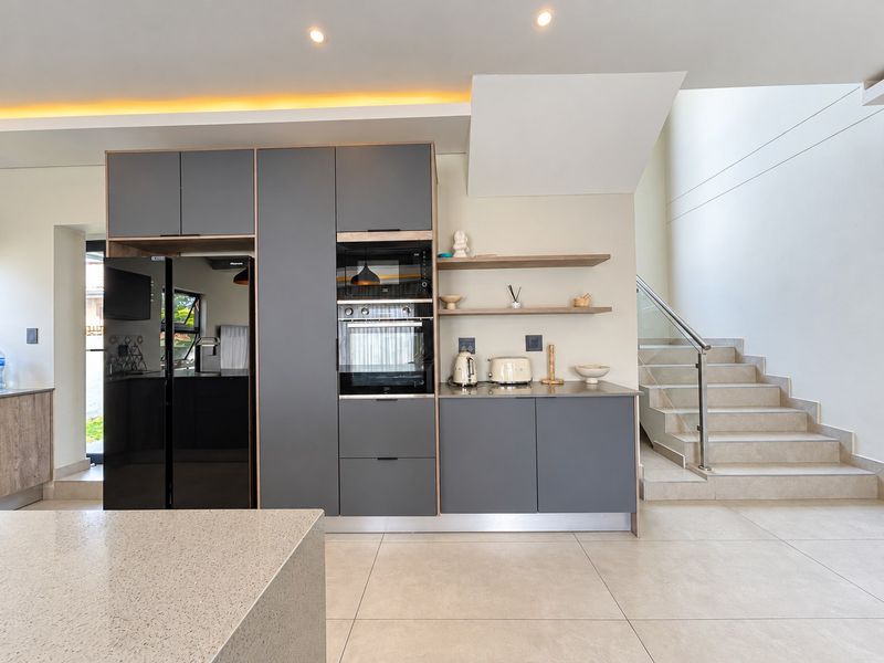Modern kitchen featuring sleek grey cabinetry, built-in appliances, stone countertops, and an adjacent staircase with a glass balustrade.