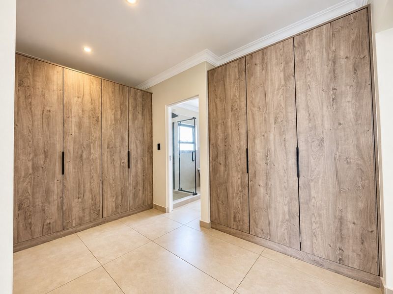 A spacious bedroom featuring extensive built-in wooden cabinetry, neutral tiled flooring, and recessed lighting, with an adjoining en-suite bathroom visible.
