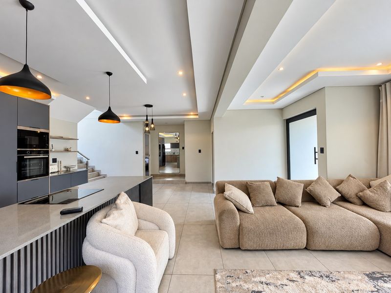 Modern open-plan living area featuring a sleek kitchen with built-in appliances, stone island, recessed ceiling lighting, and neutral tiled flooring throughout.