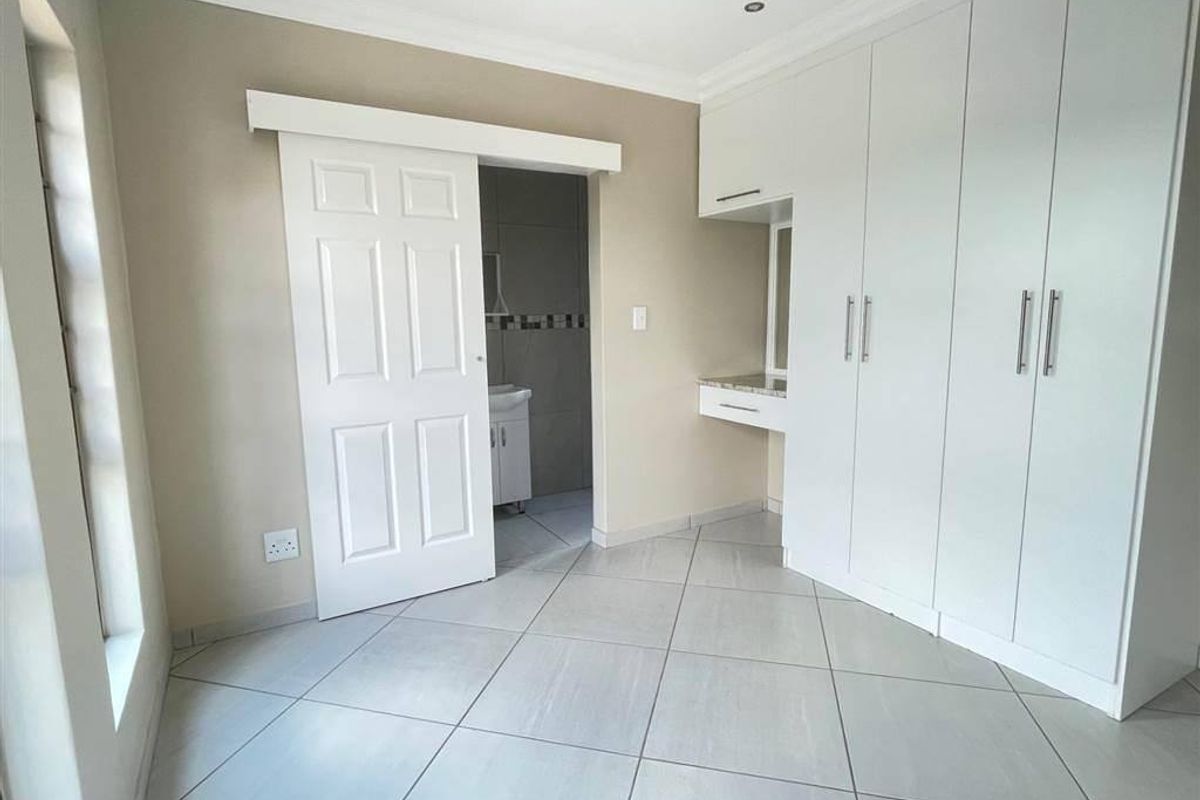 A bright, empty bedroom featuring neutral tiled flooring, extensive built-in white cabinetry with an integrated vanity, and a sliding door leading to an en-suite bathroom.
