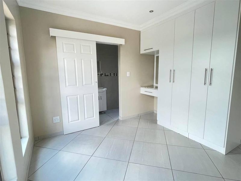 A bright, empty bedroom featuring neutral tiled flooring, extensive built-in white cabinetry with an integrated vanity, and a sliding door leading to an en-suite bathroom.