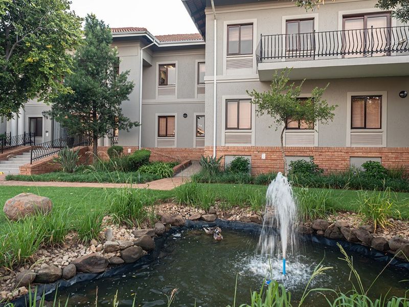 A well-maintained commercial building exterior featuring a landscaped garden, a decorative water fountain, and a balcony with wrought iron railings.