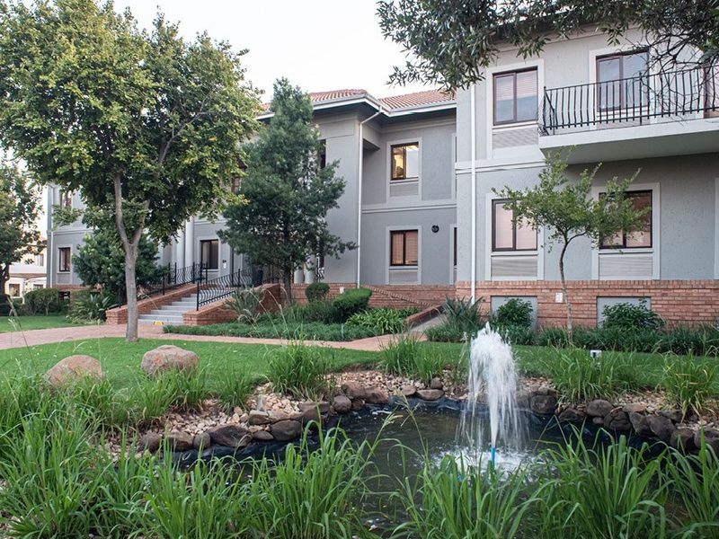 A well-maintained commercial building exterior featuring a landscaped garden, a decorative water fountain, and paved walkways leading to the building entrance.