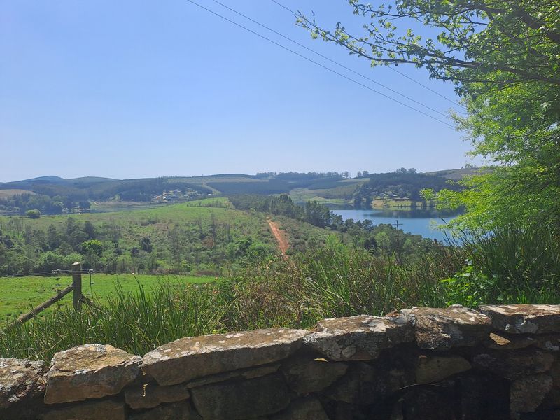 Scenic landscape view overlooking a lake, rolling hills, and natural vegetation, framed by a rustic stone retaining wall in the foreground.
