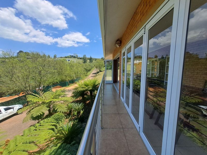 A spacious covered balcony featuring tiled flooring, a modern stainless steel balustrade, and large glass sliding doors overlooking the garden and surrounding landscape.