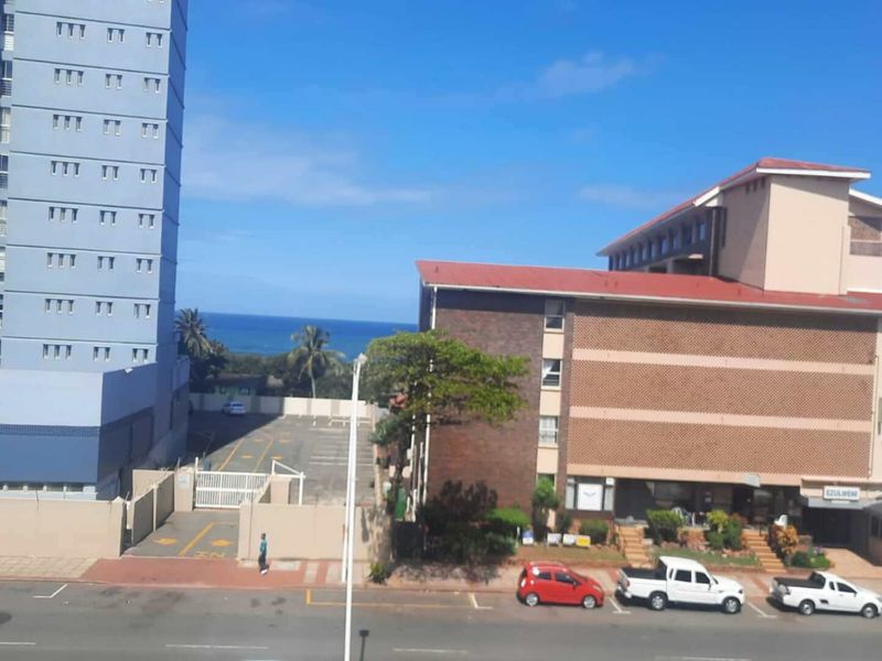 An elevated view overlooking a street, parking area, and neighbouring apartment buildings with a glimpse of the ocean in the distance.