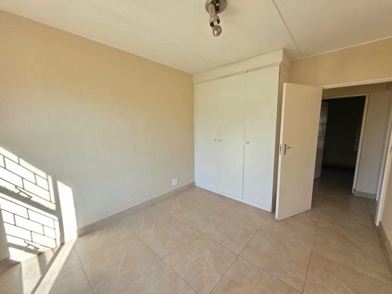 A bright, empty bedroom featuring neutral tiled flooring, built-in cupboards, and security bars on the window.
