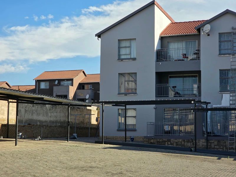 Rear view of a multi-story apartment complex featuring balconies, sliding doors, and a paved parking area with carports.