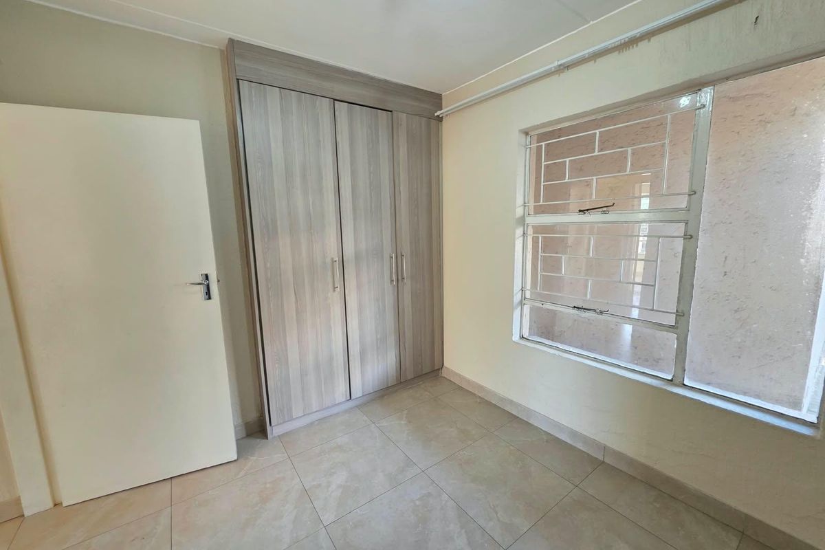A bright, empty bedroom featuring modern built-in cupboards, neutral tiled flooring, and a window fitted with security bars.