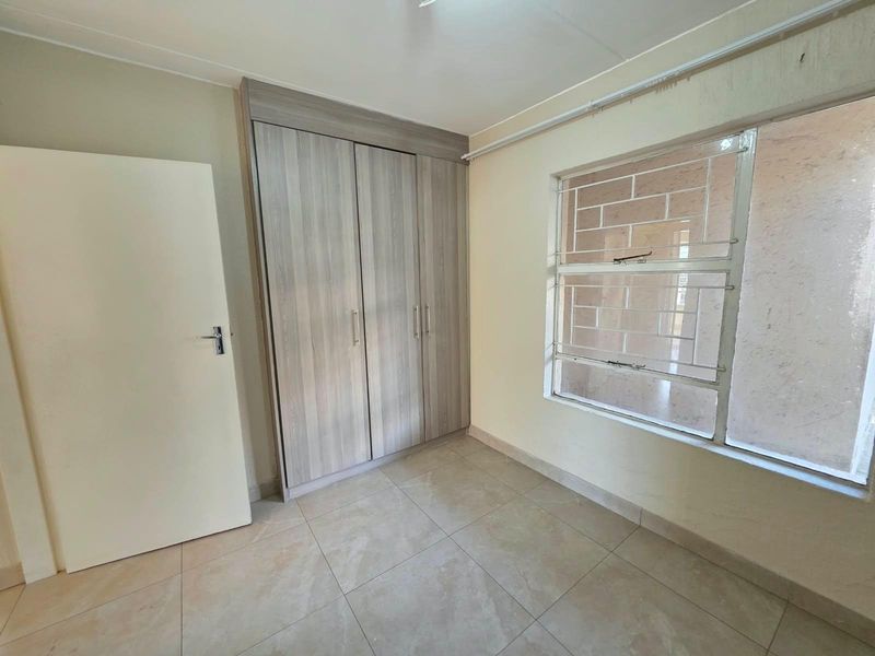 A bright, empty bedroom featuring modern built-in cupboards, neutral tiled flooring, and a window fitted with security bars.