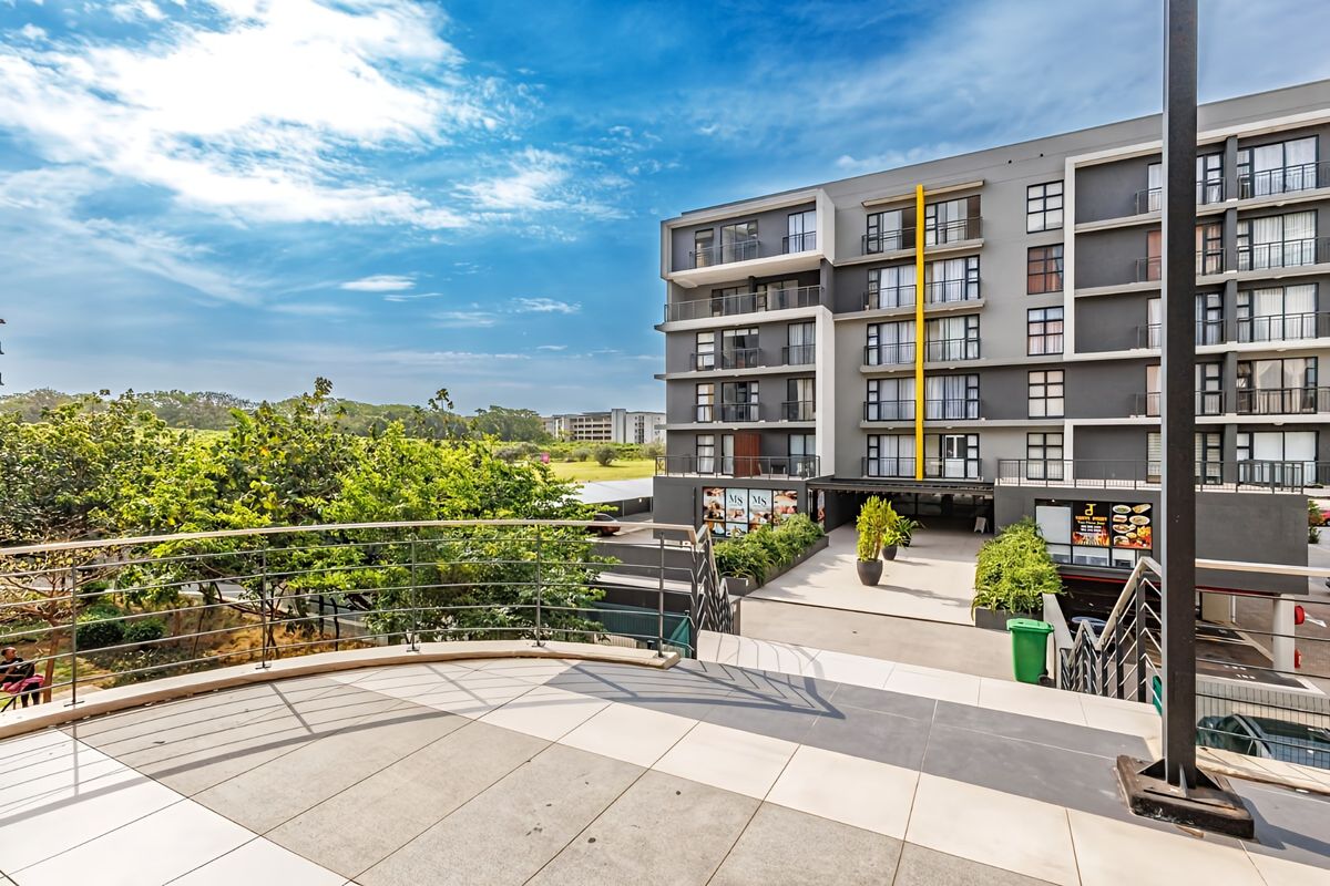 A view from a tiled balcony overlooking the rear facade of a modern apartment complex with landscaped grounds and underground parking access.