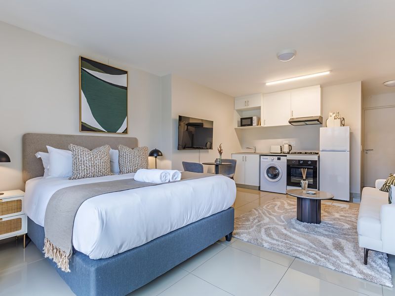 A modern open-plan studio apartment featuring a compact kitchen with built-in cabinetry, integrated appliances, and tiled flooring throughout.
