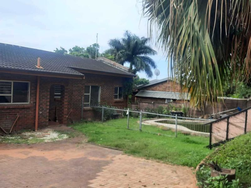 Rear view of a brick house featuring a swimming pool, paved driveway, and garden area, showing potential for landscaping and exterior maintenance.