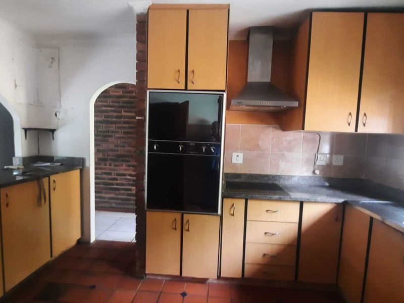 A dated kitchen featuring laminate cabinetry, granite countertops, built-in oven, and extractor fan, with tiled flooring and a brick-accented archway leading to an adjacent room.