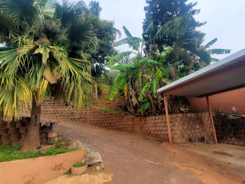 A sloped driveway featuring a brick retaining wall, mature garden landscaping, and a covered carport structure.