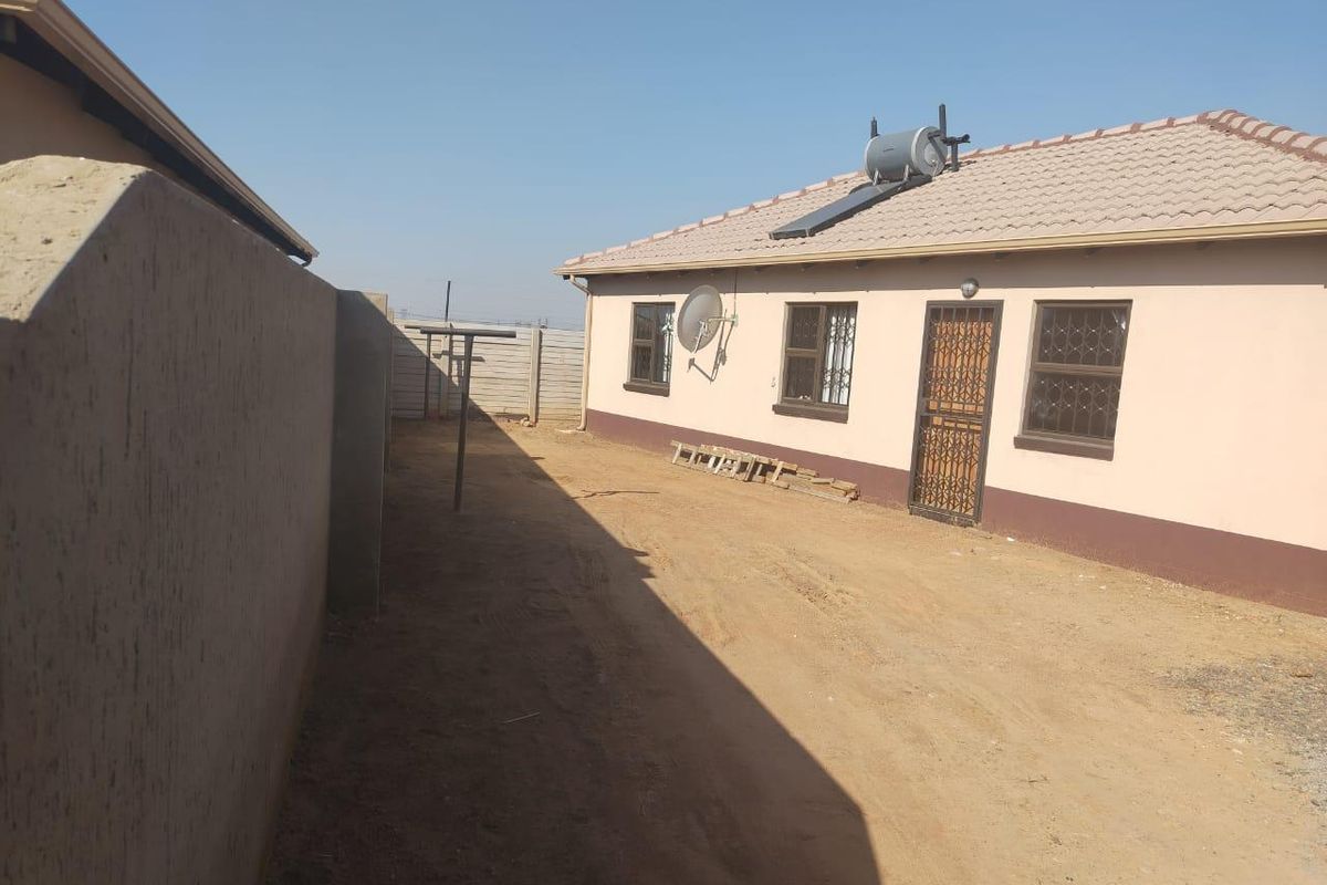 Rear exterior view of the house featuring a solar water heater, security gates, and a large unpaved yard area enclosed by a boundary wall.