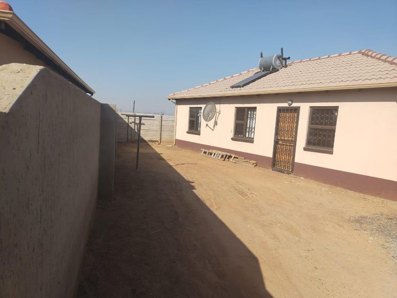 Rear exterior view of the house featuring a solar water heater, security gates, and a large unpaved yard area enclosed by a boundary wall.