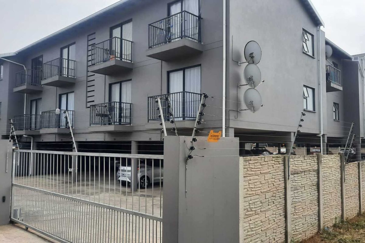 Exterior view of a multi-story apartment complex featuring balconies, secure gated access, electric fencing, and covered ground-level parking.