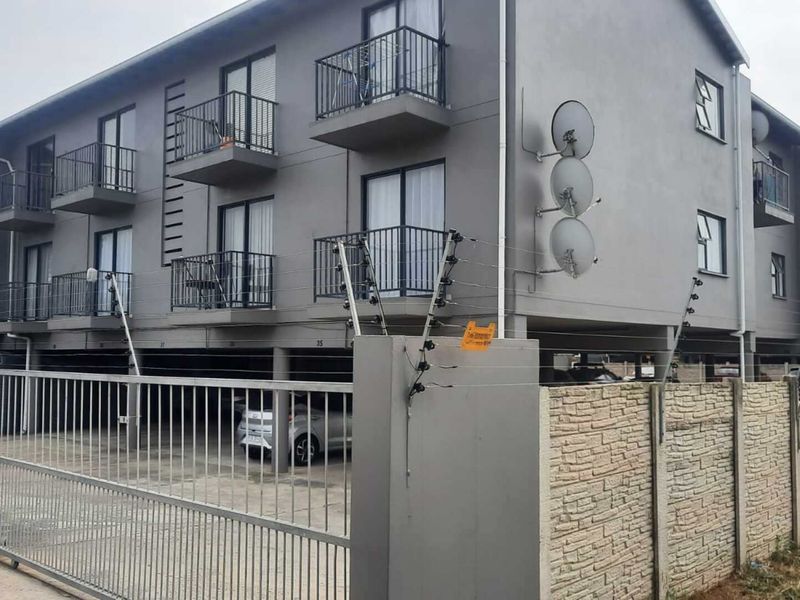 Exterior view of a multi-story apartment complex featuring balconies, secure gated access, electric fencing, and covered ground-level parking.