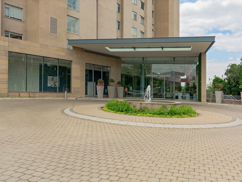 A modern apartment building entrance featuring a paved circular driveway, a central water fountain, and a prominent glass-fronted lobby with a covered canopy.