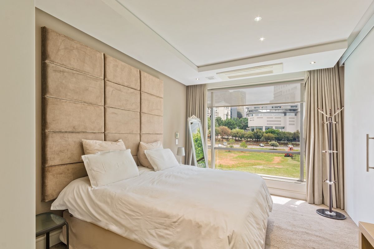 A well-maintained bedroom featuring a large upholstered headboard, recessed ceiling lighting, ducted air conditioning, and expansive windows offering pleasant urban views.