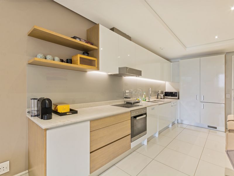 Modern kitchen featuring sleek white and wood-tone cabinetry, stone countertops, integrated appliances, and under-cabinet lighting, finished with polished floor tiles.