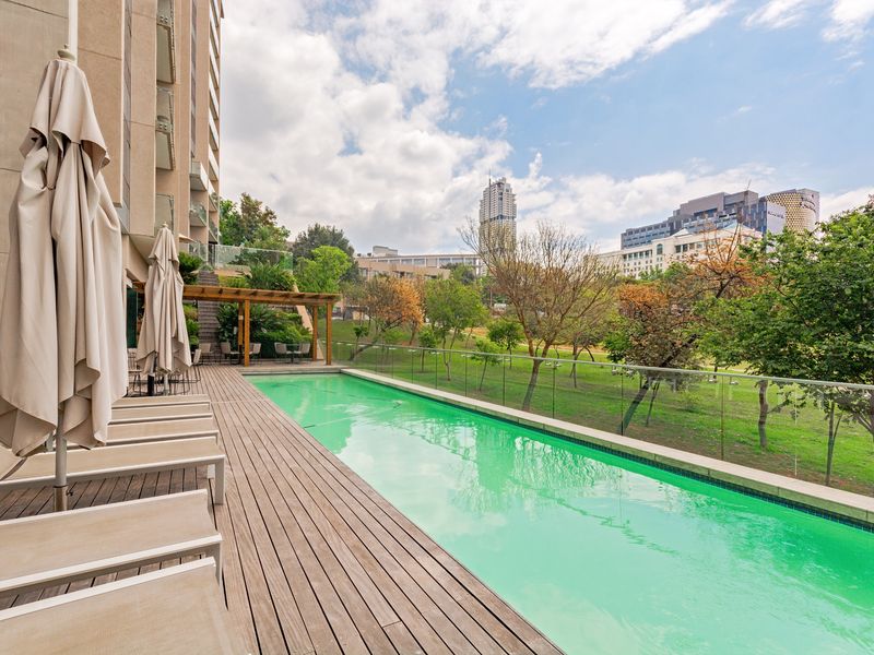 A well-maintained outdoor swimming pool area featuring timber decking, a glass balustrade, and a wooden pergola, set against a landscaped garden backdrop.
