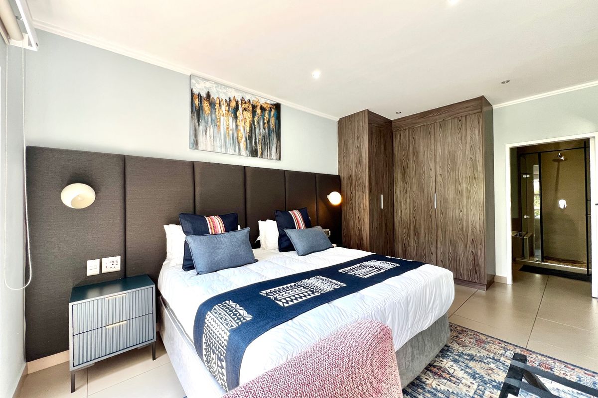 A well-maintained bedroom featuring built-in wooden cupboards, an integrated upholstered headboard, recessed lighting, and tiled flooring with an en-suite bathroom access.