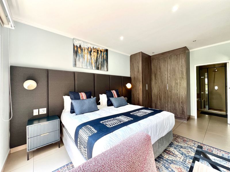 A well-maintained bedroom featuring built-in wooden cupboards, an integrated upholstered headboard, recessed lighting, and tiled flooring with an en-suite bathroom access.