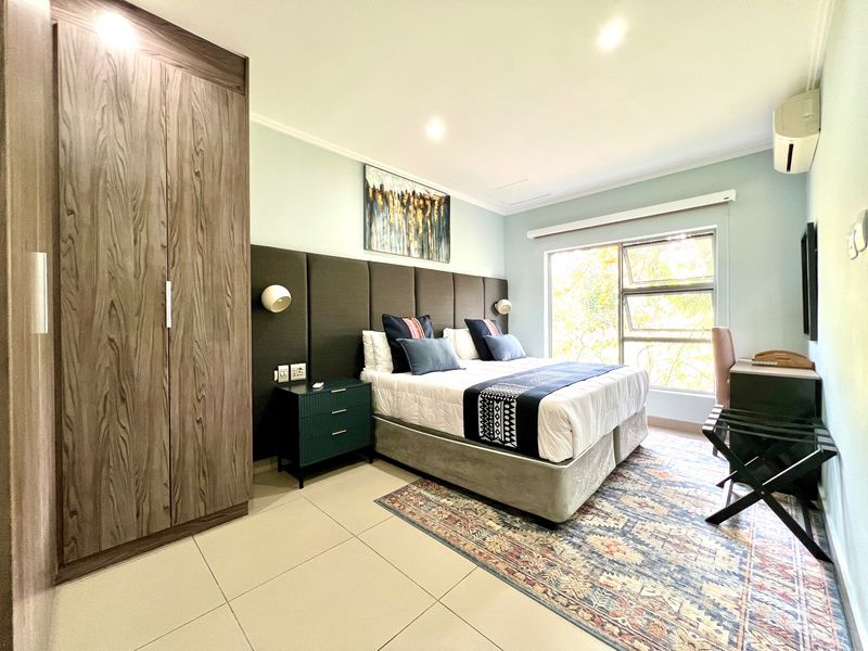 A well-maintained bedroom featuring a built-in wardrobe, upholstered headboard, tiled flooring, and a wall-mounted air conditioning unit with natural light.