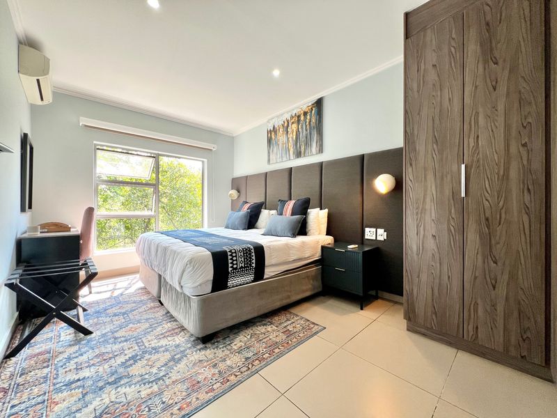 A well-maintained bedroom featuring a large built-in wardrobe, upholstered headboard, tiled flooring, and a wall-mounted air conditioning unit with natural light.