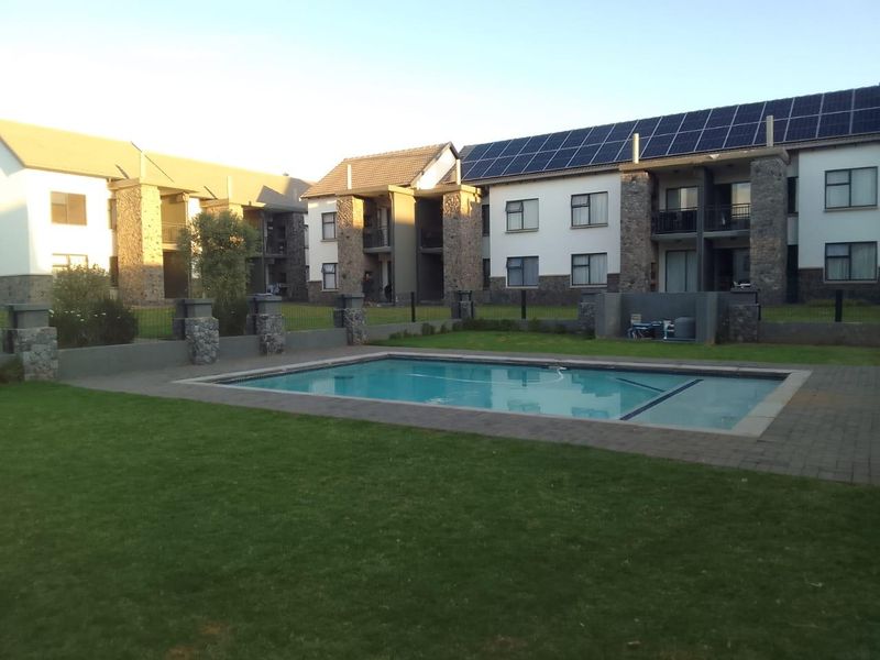 Communal swimming pool area set within a well-maintained garden, featuring stone-clad apartment building facades and roof-mounted solar panels.
