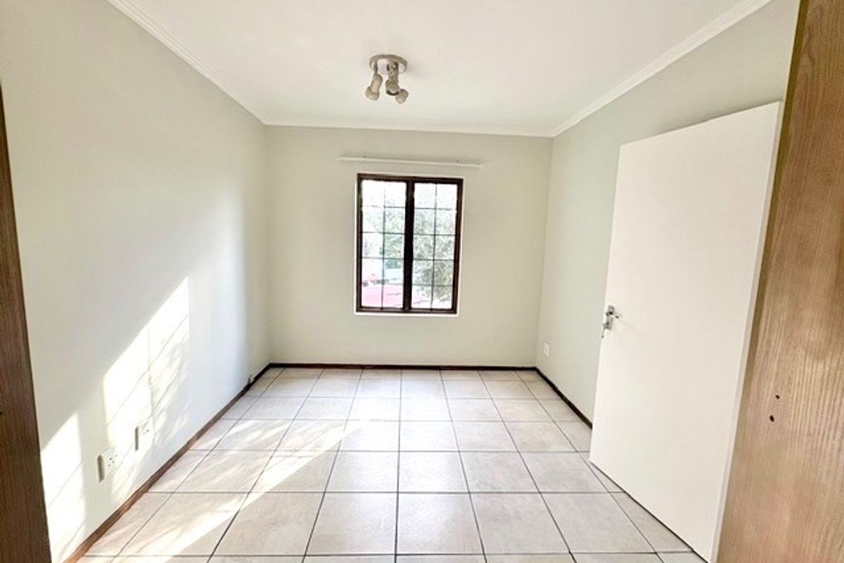 A bright, empty bedroom featuring neutral tiled flooring, a window with security bars, and a simple ceiling light fixture.
