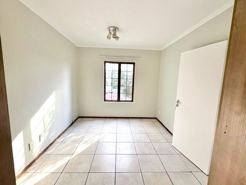 A bright, empty bedroom featuring neutral tiled flooring, a window with security bars, and a simple ceiling light fixture.