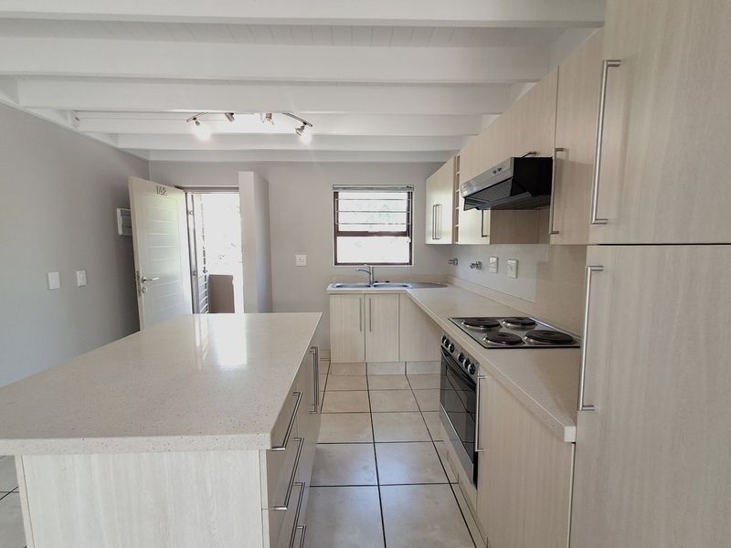 A clean, modern kitchen featuring a central island, built-in oven, electric hob, extractor fan, and ample cabinetry with tiled flooring and exposed beam ceilings.