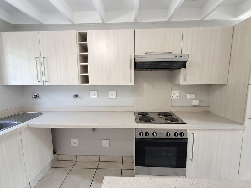 A clean, modern kitchen featuring light wood-grain cabinetry, stone countertops, integrated electric hob and oven, and a functional layout with tiled flooring.