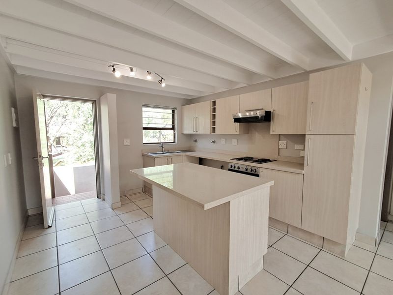 Modern kitchen featuring light wood cabinetry, stone countertops, a central island, built-in appliances, tiled flooring, and an exposed beam ceiling with direct outdoor access.
