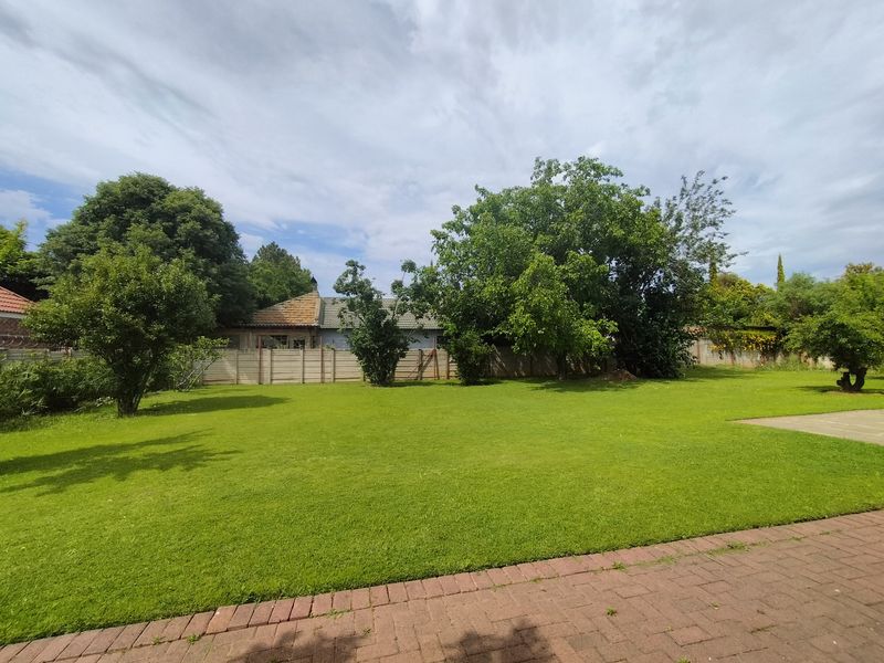 A spacious, well-maintained backyard featuring a large green lawn, mature trees, and a paved walkway, enclosed by a boundary wall.