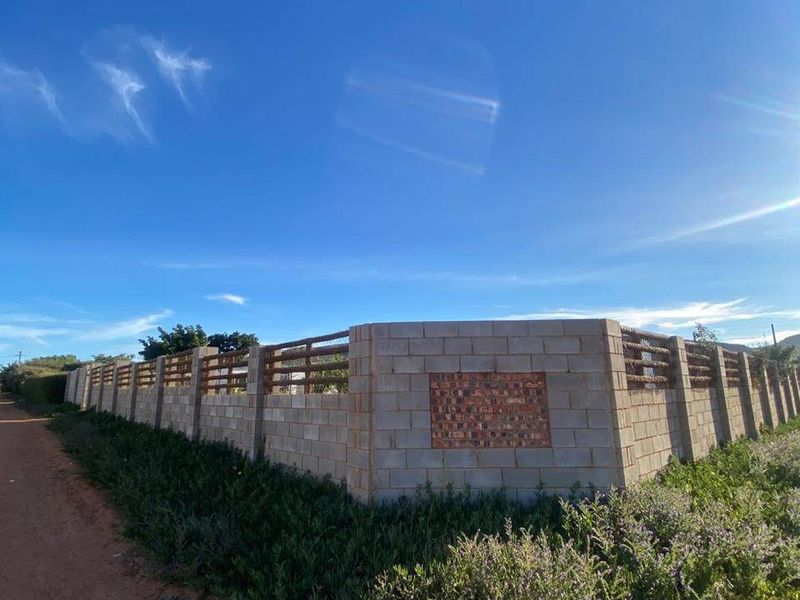 A view of a vacant residential plot featuring a newly constructed concrete block perimeter wall with timber slat detailing along an unpaved road.