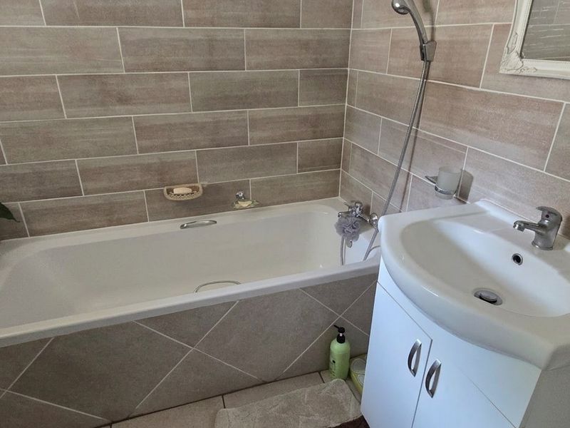A functional bathroom featuring a built-in bathtub with handheld shower attachment and a vanity unit with a modern washbasin and tiled finishes.