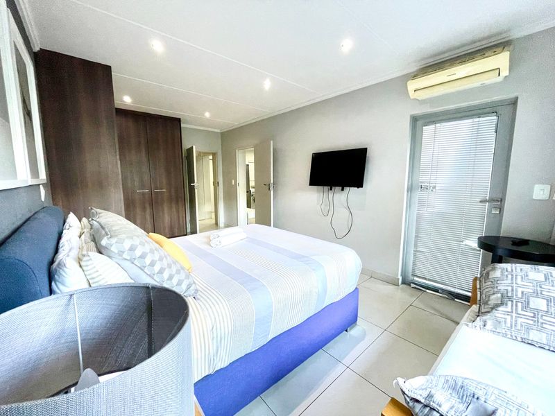 A well-maintained bedroom featuring built-in cupboards, tiled flooring, recessed lighting, and a wall-mounted air conditioning unit with direct access to an outdoor area.
