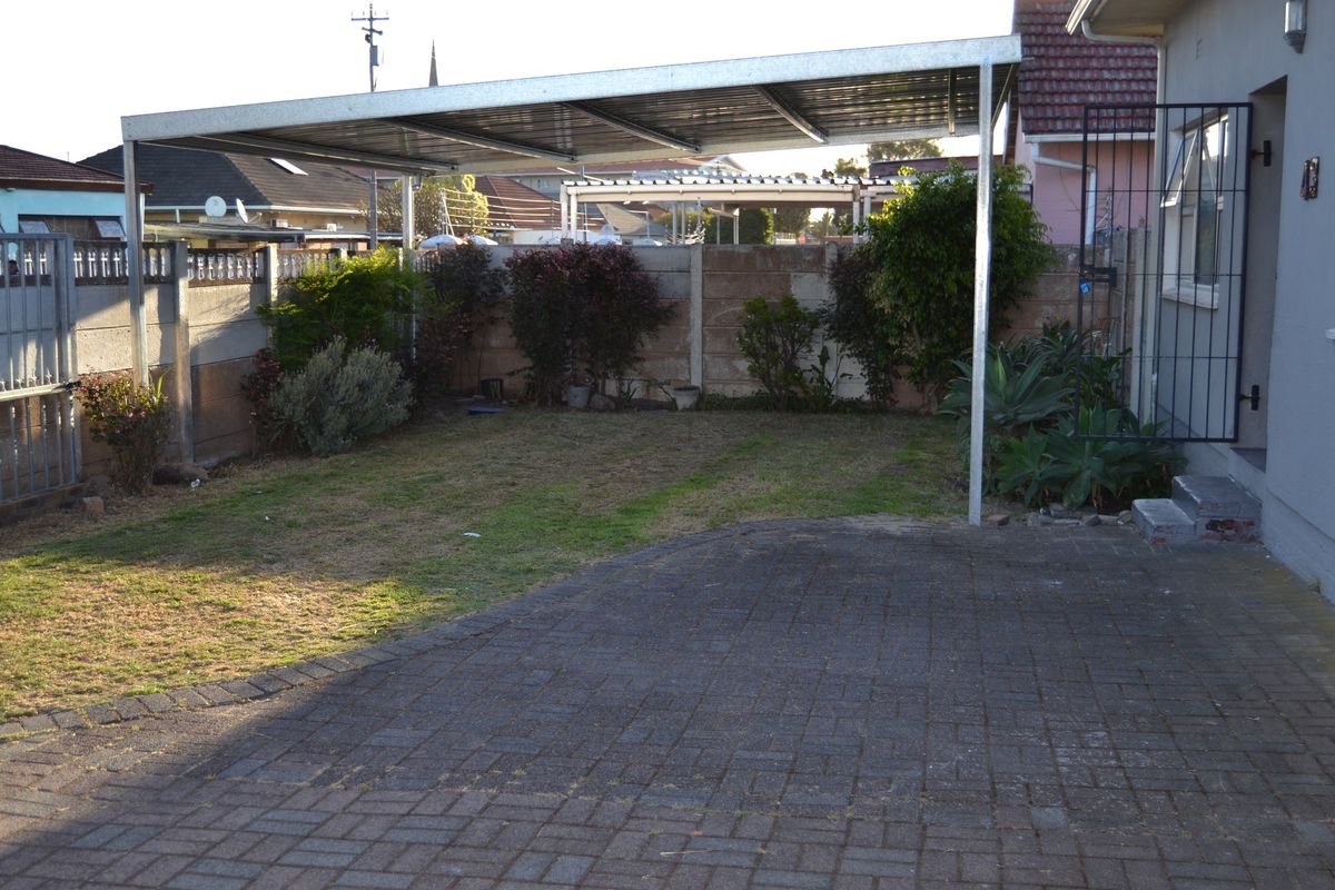 A rear exterior view featuring a paved driveway, a metal carport structure, and a precast concrete boundary wall with established garden greenery.