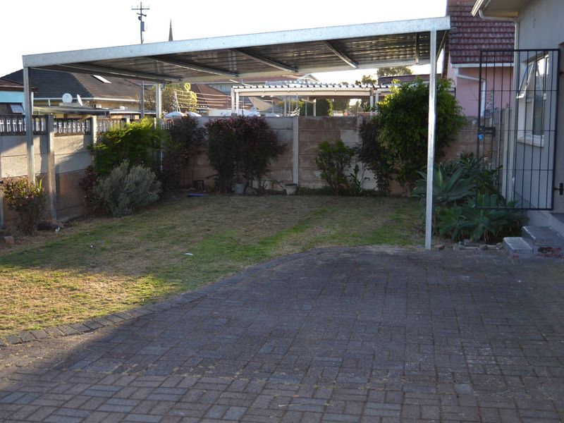A rear exterior view featuring a paved driveway, a metal carport structure, and a precast concrete boundary wall with established garden greenery.