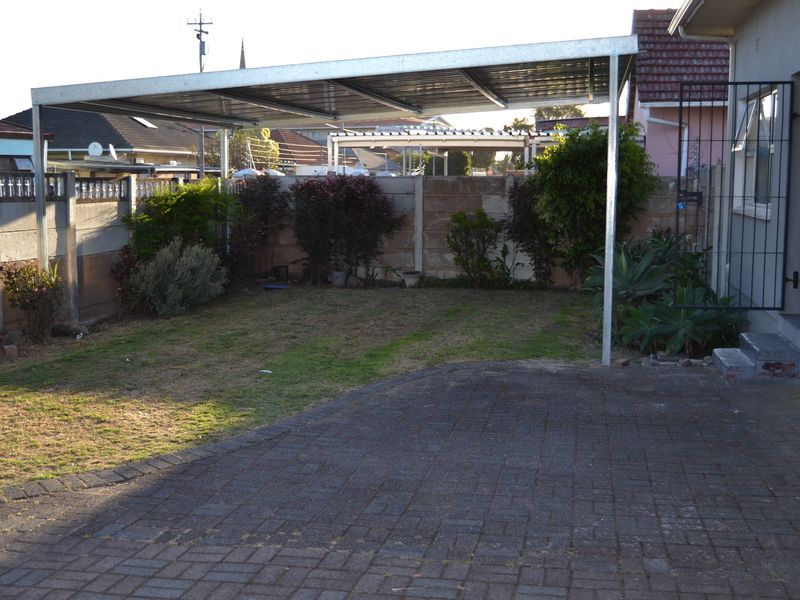 Rear exterior area featuring a paved driveway, metal carport structure, and a precast concrete boundary wall with established garden beds.