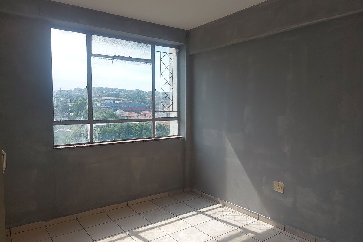 An empty bedroom featuring tiled flooring and large windows with views, currently requiring wall finishing and general maintenance to reach its full potential.