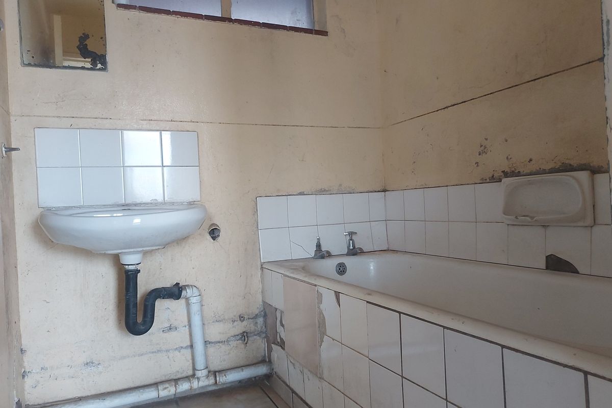 A dated bathroom requiring significant renovation, featuring a wall-mounted basin and built-in bathtub with damaged tiling and worn wall finishes.