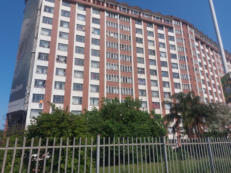 A multi-story apartment building exterior featuring a brick facade, numerous windows, and perimeter security fencing.