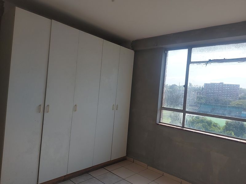 An empty bedroom featuring built-in cupboards, tiled flooring, and a large window with views. The room requires finishing touches to the wall plaster.